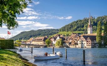 Scenic view of a historic village on Lake Constance with traditional buildings, a white church spire, and boats moored along the waterfront.