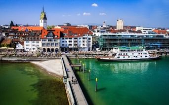 Aerial view of Friedrichshafen harbor with a ferry boat docked at the pier, green lake water, promenade with people, and historic buildings with a church tower.