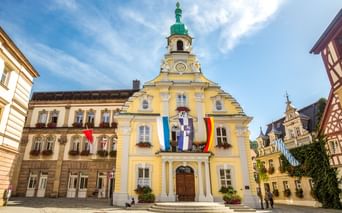 Gelbes barockes Rathaus in Kulmbach mit grünem Turm und Uhr. Bayerische und deutsche Flaggen hängen an der Fassade. Historische Gebäude rahmen den Platz.