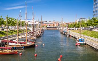 Traditionelle hölzerne Segelboote liegen in einem Kanal im Kieler Hafen unter blauem Himmel, moderne Gebäude und grüne Bäume säumen das Ufer.