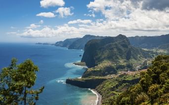 Dramatische Küstenklippen bei Funchal, Madeira, mit grüner Vegetation, türkisfarbenem Ozean und kleinen Stränden unter blauem Himmel mit Wolken.