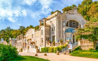 Eleganter Jugendstil-Pavillon im Kurpark Baden bei Wien mit verzierter Fassade, Bogenfenstern und großer Treppe, umgeben von grünen Rasenflächen.
