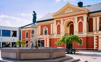 Historisches Klaipeda Theater in Korallen- und Cremefarben mit klassischer Architektur. Eine Bronzestatue steht auf einem Brunnen auf dem Platz.