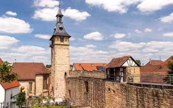 Stone tower with pointed roof and clock rising above old town wall in Marbach. Half-timbered houses with red tile roofs visible behind the fortification.