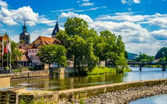 Blick auf Hamelns Altstadt entlang der Weser mit historischen Gebäuden, Kirchtürmen und grünen Bäumen unter blauem Himmel mit weißen Wolken.