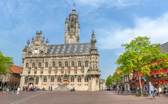 Gotisches Rathaus von Middelburg mit verzierter Fassade und hohem Turm auf einem Platz. Menschen und Radfahrer davor, flankiert von grünen Bäumen.