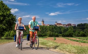 Zwei Radfahrer mit roten Rädern auf einem asphaltierten Weg durch Felder, mit Bad Wimpfens historischer Skyline und Kirchtürmen im Hintergrund.