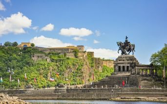 Equestrian monument at Deutsches Eck in Koblenz with fortress Ehrenbreitstein on hillside. International flags line the riverside promenade.