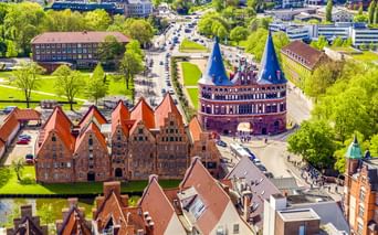 Luftaufnahme des Lübecker Holstentors mit Zwillingstürmen und roten Backsteingebäuden. Grüne Parks und Stadtstraßen umgeben das Wahrzeichen.