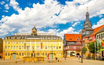 Marktplatz in Eisenach mit gelbem Rathaus, Fachwerkhäusern und Kirchturm mit Uhr unter blauem Himmel mit weißen Wolken.