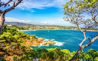 Türkisfarbene Bucht von Sant Feliu de Guíxols, eingerahmt von Pinien. Felsige Küste mit grüner Vegetation, Sandstrand und Stadt am Wasser.