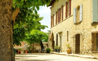 Traditionelles Steinhaus bei Uzès mit Bogentüren und Fensterläden. Ein großer Baum spendet Schatten über einem sonnigen Innenhof mit Topfpflanzen.