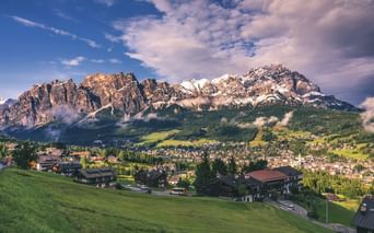 Alpendorf in Südtirol mit traditionellen Häusern auf grünen Wiesen. Dramatische Dolomitengipfel erheben sich hinter Wäldern unter blauem Himmel.