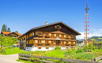 Traditionelles bayerisches Holzhaus mit Blumenkästen in Oberstaufen. Ein dekorativer Maibaum mit Fahnen steht neben dem Gebäude auf grüner Wiese.