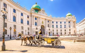 Gelber Fiaker vor der Alten Hofburg in Wien mit weißer Barockfassade und grünen Kupferkuppeln unter blauem Himmel.