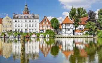 Historischer Hafen von Glückstadt mit bunten Gebäuden, die sich im ruhigen Wasser spiegeln. Weißes Gebäude mit Turm, Fachwerkhäuser und Boote am Kai.