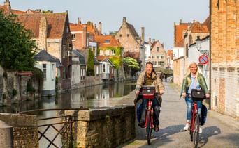 Zwei Radfahrer auf roten Fahrrädern auf einer Kopfsteinpflasterstraße neben einem Kanal in Flandern, mit historischen Backsteingebäuden und orangefarbenen Ziegeldächern.
