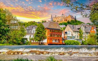 Marburg cityscape with colorful historic buildings along a weir on the Lahn River. Marburg Castle sits on the hilltop under a dramatic sunset sky.