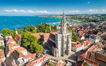 Luftaufnahme des Konstanzer Münsters mit gotischem Turm über den roten Dächern der Altstadt und dem Bodensee mit Segelbooten unter blauem Himmel.