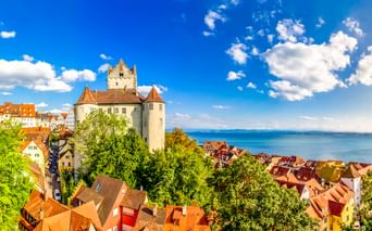 Burg Meersburg mit weißen Mauern und Turm über rotgedeckten Häusern, mit Blick auf den Bodensee unter blauem Himmel mit weißen Wolken.