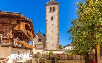 Stone bell tower of Innichen Collegiate Church rising against blue sky, with traditional Alpine wooden house and green tree framing the scene.