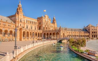 Plaza de España in Sevilla zeigt verzierte Backsteingebäude mit Türmen und Bögen entlang eines Kanals mit Brücke. Besucher flanieren über den Platz.