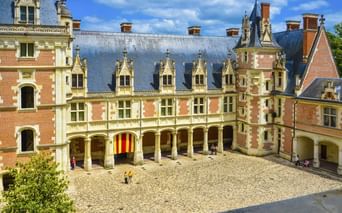 Kopfsteinpflaster-Innenhof des Château de Blois mit Renaissance-Architektur, verzierten Dachgauben und Arkadengang unter blauem Himmel.