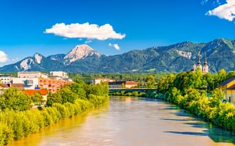 View of Villach with the Drau River flowing through green banks, colorful buildings, a church, and dramatic mountain peaks under blue sky.