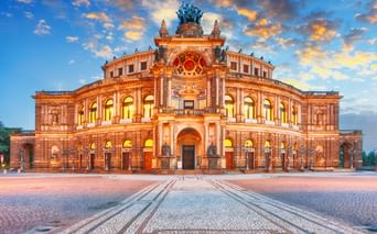 Beleuchtete Semperoper in Dresden mit prachtvoller Barockarchitektur unter dramatischem Sonnenuntergangshimmel mit orangen und blauen Wolken.