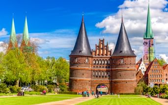 Das Holstentor, ein mittelalterliches Stadttor aus rotem Backstein mit zwei runden Türmen in Lübeck, umgeben von grünen Rasenflächen und Kirchtürmen.