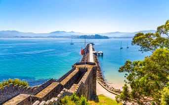 Steinmauern der Festung Monterreal erstrecken sich in die Bucht von Baiona mit Segelbooten im türkisfarbenen Wasser und Bergen in der Ferne.