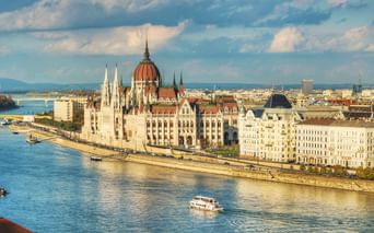 Panoramablick auf das ungarische Parlamentsgebäude am Donauufer in Budapest mit Booten auf dem Wasser und einer Brücke im Hintergrund.