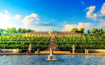 Schloss Sanssouci in Potsdam mit gelber Fassade und terrassierten Weingärten. Brunnen im Vordergrund, Besucher auf Treppe, blauer Himmel mit Wolken.