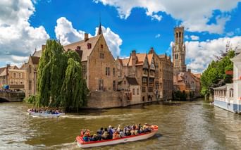 Zwei Touristenboote auf einem Kanal in Brügge mit mittelalterlichen Gebäuden und dem Belfried-Turm unter blauem Himmel mit weißen Wolken.