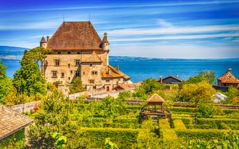 Mittelalterliches Schloss in Yvoire mit Steinmauern und rotem Ziegeldach mit Blick auf den Genfersee. Formaler Garten mit geometrischen Hecken im Vordergrund.