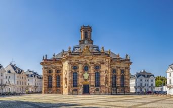 Ludwigskirche, eine barocke Kirche mit gelber Sandsteinfassade und Turm, auf einem Kopfsteinpflasterplatz in Saarbrücken unter blauem Himmel.