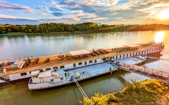 MS Prinzessin Katharina Flusskreuzfahrtschiff an der Donau bei Sonnenuntergang. Das weiße Schiff hat ein Holzdeck mit Fahrrädern und Liegestühlen.