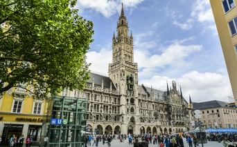Marienplatz in München mit dem gotischen Rathausturm vor blauem Himmel. Fußgänger versammeln sich auf dem Platz mit grünen Bäumen links.