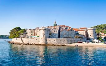 Blick auf die Altstadt von Korcula mit steinernen Befestigungsmauern, historischen Gebäuden mit Terrakottadächern und Kirchturm unter blauem Himmel.