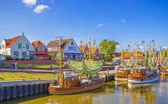 Traditionelle Fischerboote mit Netzen liegen im Hafen von Greetsiel. Bunte Häuser mit roten Dächern säumen das Ufer unter blauem Himmel.