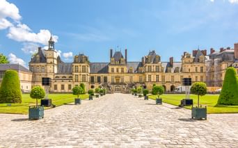 Fassade des Schlosses Fontainebleau mit Kopfsteinpflaster-Innenhof, gepflegten Rasenflächen und Formschnittbäumen unter blauem Himmel.