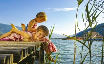 Zwei Kinder in passenden Badeanzügen spielen auf einem Holzsteg an einem See. Berge und Schilf sind im Hintergrund unter blauem Himmel sichtbar.
