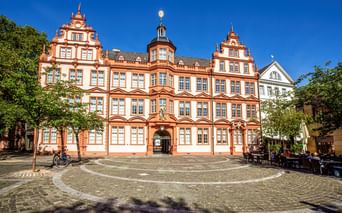 Rot-weißes Gutenberg-Museum in Mainz im Renaissance-Stil mit verzierten Giebeln und zentralem Turm an einem Kopfsteinpflasterplatz.