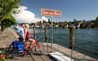 Zwei Radfahrer mit rotem Fahrrad am Ortsschild Stein am Rhein am Rheinufer. Historische Stadtgebäude sichtbar über dem türkisfarbenen Wasser.