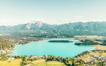 Luftaufnahme vom türkisfarbenen Faaker See in Österreich mit bewaldeten Inseln, grünen Wiesen und Bergkette im Hintergrund unter klarem Himmel.