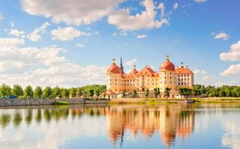 Moritzburg Castle with yellow facade and red domes reflected in surrounding lake under blue sky with white clouds.