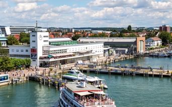 Fahrgastschiff im Friedrichshafener Hafen am Bodensee. Weiße Hafengebäude und Holzstege unter blauem Himmel mit weißen Wolken.