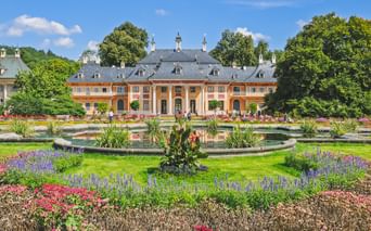 Schloss Pillnitz mit oranger Fassade und grauem Dach hinter Zierbrunnen und bunten Blumenbeeten im barocken Garten unter blauem Himmel.