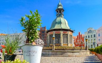 Verzierter Pavillon mit Kupferkuppel auf Kopfsteinpflasterplatz in Wismar. Bunte historische Gebäude im Hintergrund unter blauem Himmel.