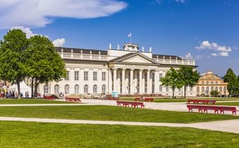 The neoclassical Fridericianum building in Kassel with columned facade, viewed across a green lawn with red benches under blue sky.